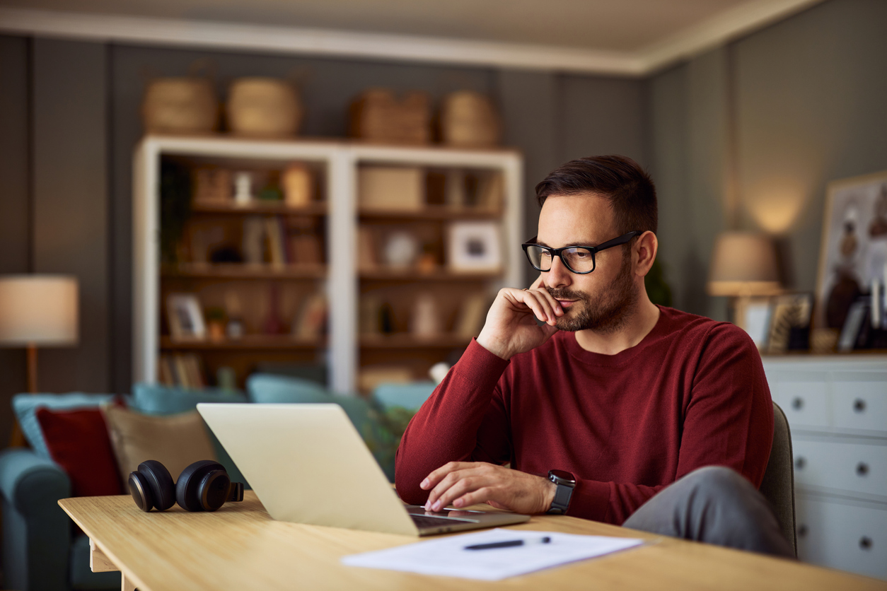 A man wearing glasses, thinking at his desk and working on his laptop.